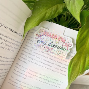 Puede incluir: Una pegatina holográfica rosa y blanca con el texto "Hello I'm very demure" en una pancarta rosa con flores blancas y un lazo rosa. La pegatina tiene un acabado brillante.