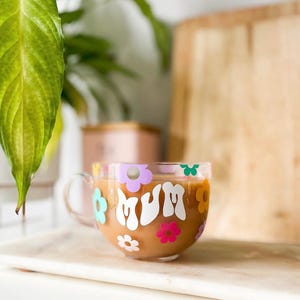 May include: A clear glass mug with a brown beverage inside. The mug has a white "MUM" design with colourful flowers around it.