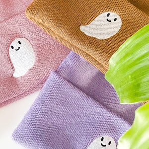 Puede incluir: Tres gorros de punto en diferentes colores: rosa, marrón y morado. Cada gorro tiene un fantasma bordado blanco con una cara sonriente.