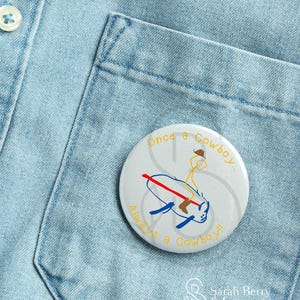 May include: A white button with the text "Once a Cowboy... Always a Cowboy!!" in yellow and blue. The button features a cartoon cowboy riding a blue object with a red line through it. The button is on a light blue denim shirt.