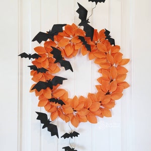 Op de afbeelding: Een Halloweenkrans gemaakt van oranje viltbladeren met zwarte viltvleermuizen die eraan hangen. De krans hangt aan een witte deur.