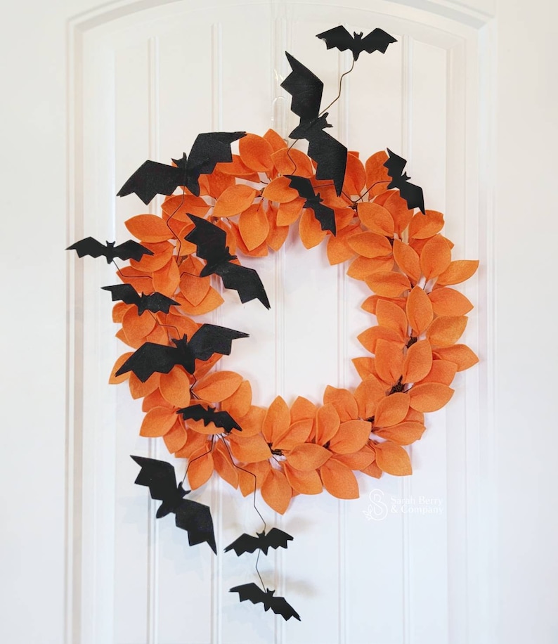 Op de afbeelding: Een Halloweenkrans gemaakt van oranje viltbladeren met zwarte viltvleermuizen die eraan hangen. De krans hangt aan een witte deur.