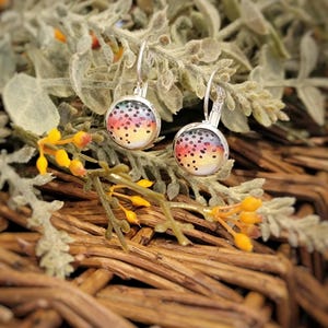 May include: Silver-toned leverback earrings with round, colorful fish scale designs. The earrings feature a vibrant pattern of red, yellow, and black dots, set against a white background. The earrings are displayed on a bed of greenery and woven wood.