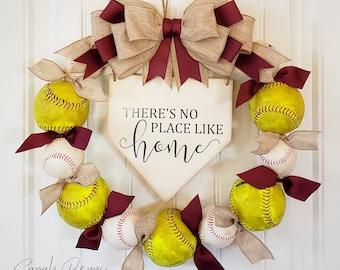 May include: A softball wreath with a burlap bow and maroon ribbons. The wreath is decorated with softballs and a wooden sign that reads "There's no place like home".