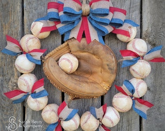 May include: A baseball-themed wreath featuring baseballs, a baseball glove, and red, white, and blue bows. The wreath is arranged on a wooden background, creating a rustic and sporty aesthetic. The baseball glove is in the center, holding a baseball.