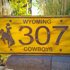 May include: Yellow wooden sign with brown text and a silhouette of a cowboy riding a horse. The sign reads "WYOMING 307 COWBOYS".