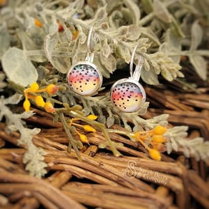 May include: Silver leverback earrings with a colorful, speckled design. The earrings feature a round, glass-covered image with a rainbow of colors, including yellow, orange, red, and black dots. The earrings are set against a backdrop of greenery and woven wood.