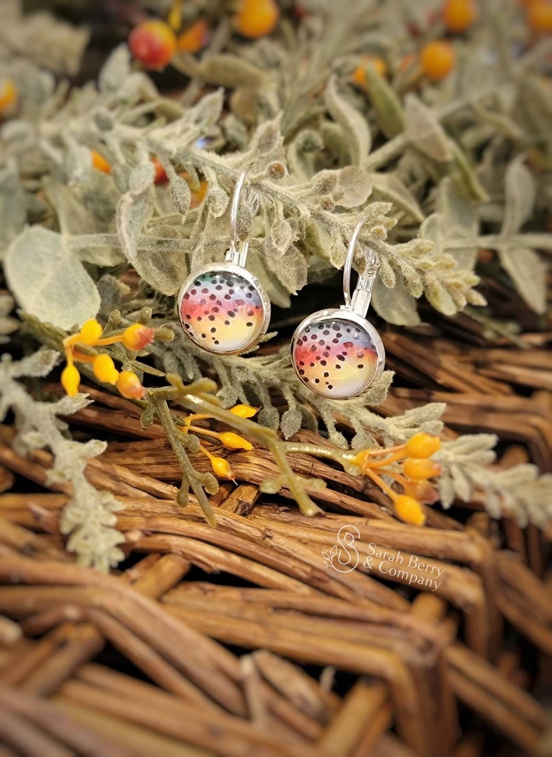 May include: Silver leverback earrings with a colorful, speckled design. The earrings feature a round, glass-covered image with a rainbow of colors, including yellow, orange, red, and black dots. The earrings are set against a backdrop of greenery and woven wood.