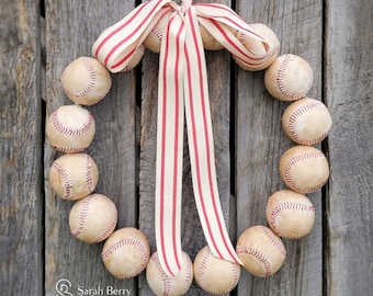 May include: A red and white striped ribbon is tied around a wreath of baseballs. The wreath is made of 12 baseballs, each with a white stitching pattern. The wreath is hanging on a wooden wall.