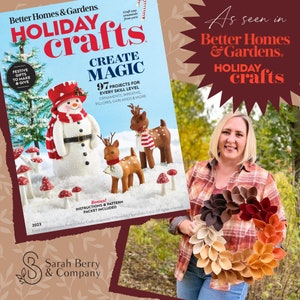 Op de afbeelding: Een tijdschriftcover voor Better Homes & Gardens Holiday Crafts met een sneeuwpop, twee herten en rode en witte paddenstoelen. De tekst op de cover luidt "Create Magic" en "97 Projects for Every Skill Level". Het tijdschrift wordt vastgehouden door een vrouw die een geruit overhemd en een spijkerbroek draagt, en een krans van viltbladeren in tinten bruin, oranje en rood vasthoudt.