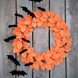 Op de afbeelding: Een Halloweenkrans gemaakt van oranje viltbladeren met zwarte vleermuizen die eromheen hangen. De krans hangt aan een houten achtergrond.