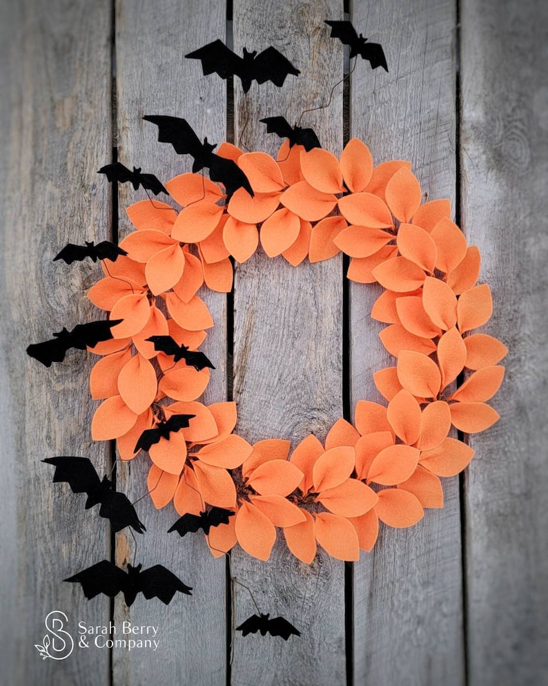 Op de afbeelding: Een Halloweenkrans gemaakt van oranje viltbladeren met zwarte vleermuizen die eromheen hangen. De krans hangt aan een houten achtergrond.