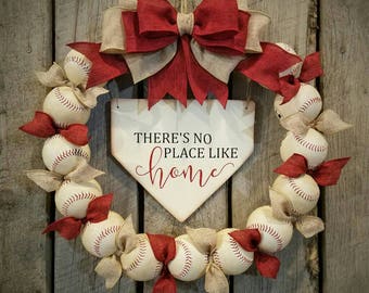 May include: A baseball themed wreath with a red and burlap bow. The wreath is decorated with baseballs and red bows. A wooden sign with the text "There's no place like home" is in the center of the wreath.