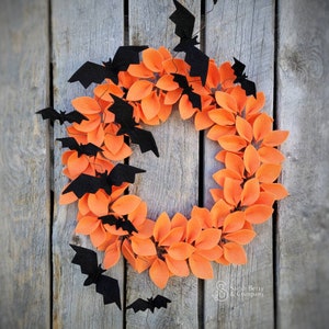 Op de afbeelding: Een Halloweenkrans gemaakt van oranje viltbladeren met zwarte viltvleermuizen die eromheen hangen. De krans hangt aan een houten achtergrond.