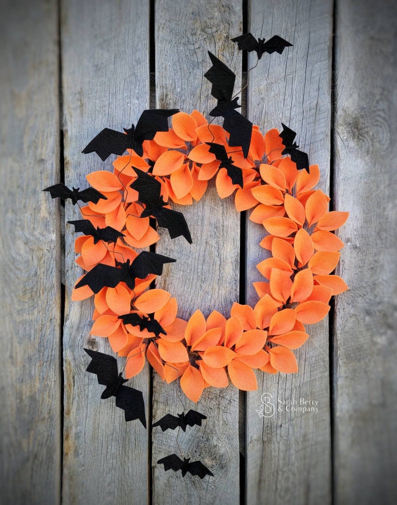 Op de afbeelding: Een Halloweenkrans gemaakt van oranje viltbladeren met zwarte viltvleermuizen die eromheen hangen. De krans hangt aan een houten achtergrond.