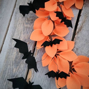 Op de afbeelding: Een Halloweenkrans gemaakt van oranje viltbladeren met zwarte vleermuizen die eraan hangen. De krans hangt aan een houten achtergrond.