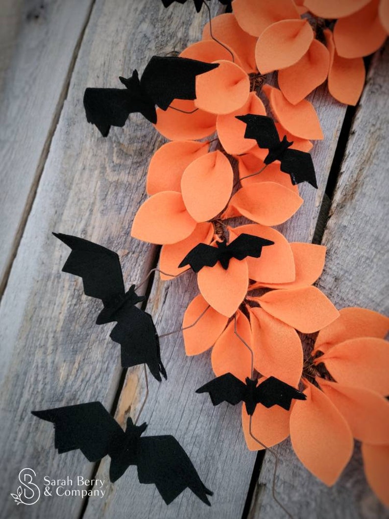 Op de afbeelding: Een Halloweenkrans gemaakt van oranje viltbladeren met zwarte vleermuizen die eraan hangen. De krans hangt aan een houten achtergrond.