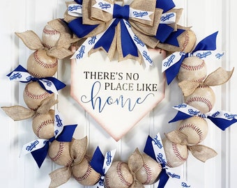 May include: A baseball-themed wreath featuring baseballs, burlap ribbon, and blue bows with "Dodgers" text. A home plate-shaped sign in the center reads "THERE'S NO PLACE LIKE home". The wreath is a decorative item for a door or wall.