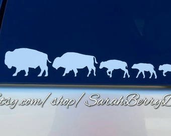May include: A white vinyl decal of a family of five bison, with the largest bison on the left and the smallest on the right. The decal is on a blue car window. The text "www.Etsy.com/shop/SarahBerryDesigns" is visible at the bottom of the image.