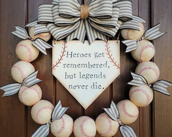 May include: A baseball themed wreath with a wooden sign that reads "Heroes get remembered, but legends never die." The wreath is decorated with baseballs, black and white striped ribbon, and a large bow.