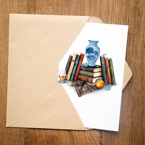 May include: A greeting card featuring an illustration of a stack of vintage books, a blue and white ceramic vase, a glass of amber liquid, a plaid fabric scrap, and an orange, all arranged together on a plain white background inside a kraft envelope.