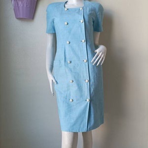 May include: A light blue dress with short sleeves and a double breasted button closure. The dress has a straight silhouette and a knee-length hem.