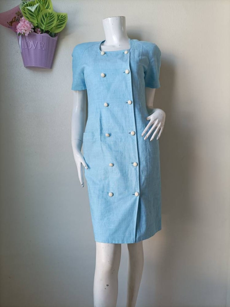 May include: A light blue dress with short sleeves and a double breasted button closure. The dress has a straight silhouette and a knee-length hem.