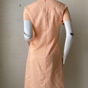 K&ouml;nnte beinhalten: Pfirsichfarbenes Kleid mit kurzen &Auml;rmeln. Das Kleid hat ein ma&szlig;geschneidertes Design mit rundem Ausschnitt und einer vertikalen Naht auf der R&uuml;ckseite. Das Kleid wird auf einer Schaufenquin ausgestellt.