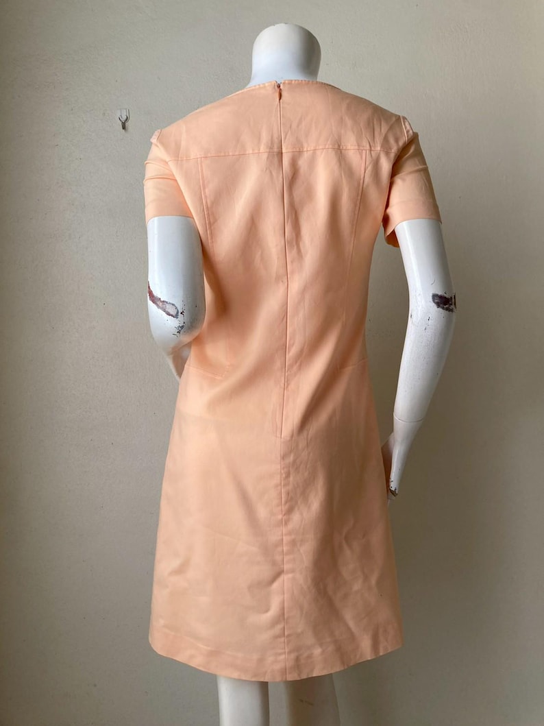 K&ouml;nnte beinhalten: Pfirsichfarbenes Kleid mit kurzen &Auml;rmeln. Das Kleid hat ein ma&szlig;geschneidertes Design mit rundem Ausschnitt und einer vertikalen Naht auf der R&uuml;ckseite. Das Kleid wird auf einer Schaufenquin ausgestellt.