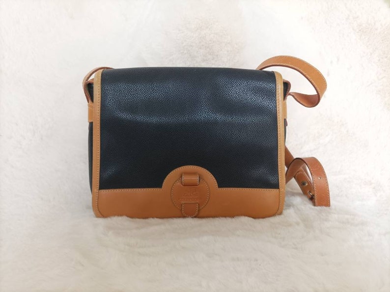 May include: A black leather satchel with a brown leather trim and a metal clasp. The bag has a long adjustable strap.