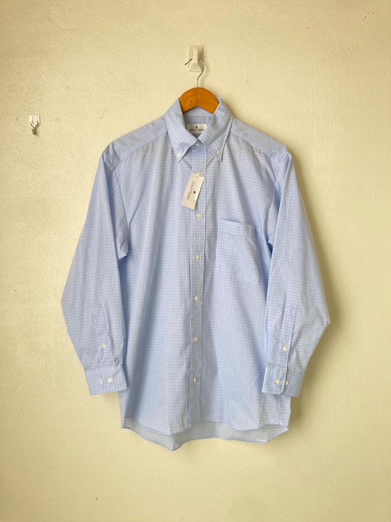 May include: A light blue long-sleeved button-down shirt with a white and light blue checkered pattern. The shirt has a pocket on the left side and a white tag with the text "Country Club Prep" on the front.