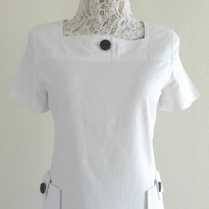 May include: White short-sleeved dress with a square neckline and two black buttons on the front.