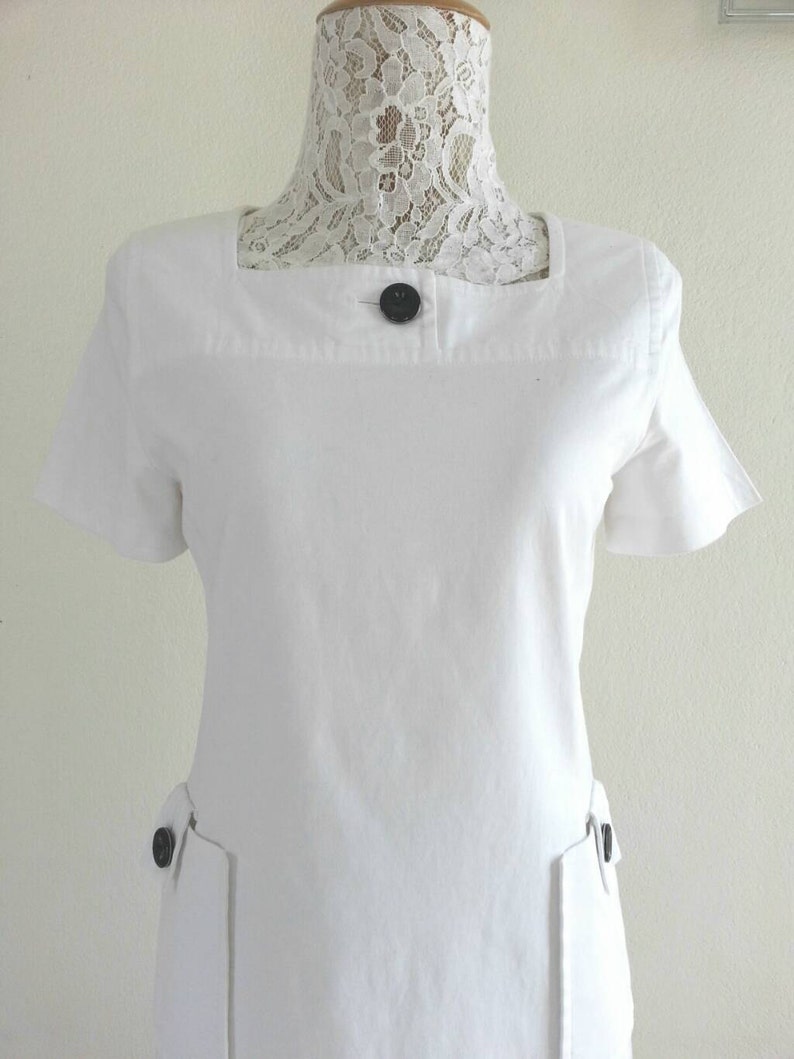 May include: White short-sleeved dress with a square neckline and two black buttons on the front.