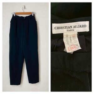 May include: Dark navy blue trousers hanging on a hanger. The trousers have a button closure and pockets. A close-up shows a tag that reads "CHRISTIAN AUJARD PARIS".
