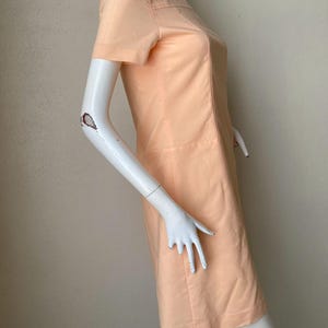 K&ouml;nnte beinhalten: Pfirsichfarbenes Kleid mit kurzen &Auml;rmeln. Das Kleid hat ein schlichtes, ma&szlig;geschneidertes Design mit rundem Ausschnitt und dezenten Nahtdetails. Das Kleid wird auf einer Schaufensterpuppe pr&auml;sentiert.