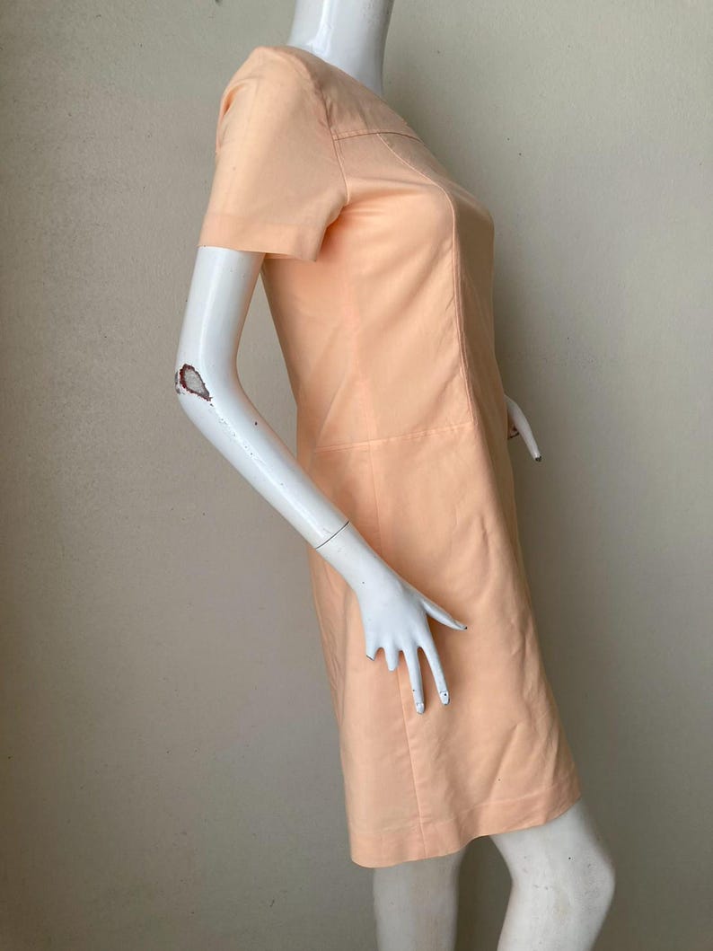 K&ouml;nnte beinhalten: Pfirsichfarbenes Kleid mit kurzen &Auml;rmeln. Das Kleid hat ein schlichtes, ma&szlig;geschneidertes Design mit rundem Ausschnitt und dezenten Nahtdetails. Das Kleid wird auf einer Schaufensterpuppe pr&auml;sentiert.