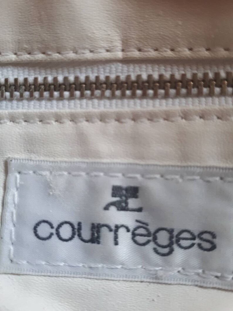 K&ouml;nnte beinhalten: Wei&szlig;e Ledertasche mit einem silbernen Rei&szlig;verschluss und einem wei&szlig;en Etikett mit dem Text "Courr&egrave;ges" in schwarzen Buchstaben.