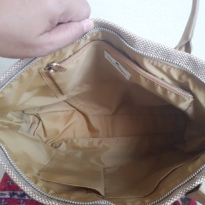 May include: Beige canvas tote bag with a zip closure and a small zipped pocket inside. The bag has a beige fabric lining.