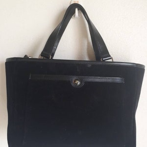 K&ouml;nnte beinhalten: Eine schwarze Cord-Tragetasche mit einem Leder-Oberhenkel und einem Klappenverschluss. Die Tasche hat eine kleine Tasche auf der Vorderseite.