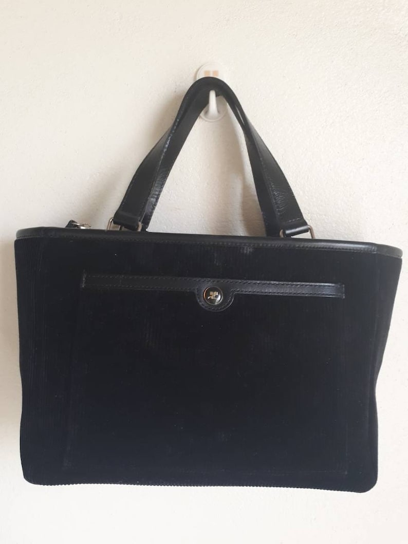 K&ouml;nnte beinhalten: Eine schwarze Cord-Tragetasche mit einem Leder-Oberhenkel und einem Klappenverschluss. Die Tasche hat eine kleine Tasche auf der Vorderseite.