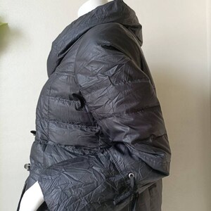 May include: A dark grey puffer coat with a high collar and a cinched waist. The coat has a quilted design and long sleeves with drawstrings at the cuffs. It is displayed on a mannequin.