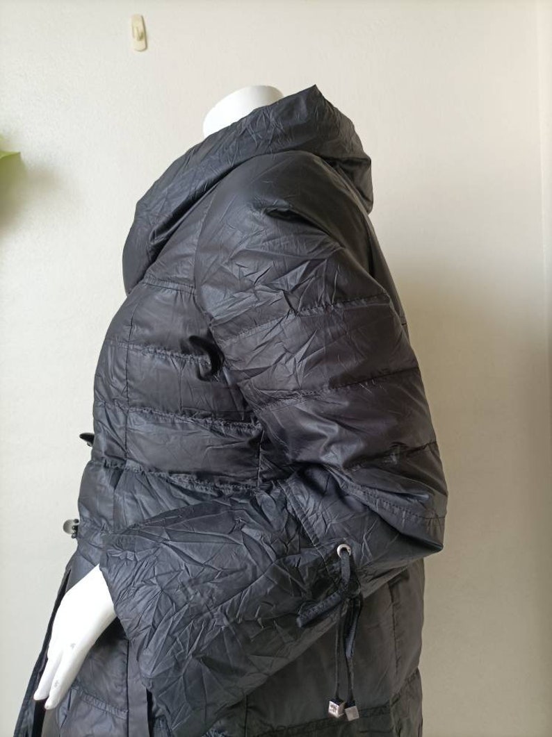May include: A dark grey puffer coat with a high collar and a cinched waist. The coat has a quilted design and long sleeves with drawstrings at the cuffs. It is displayed on a mannequin.