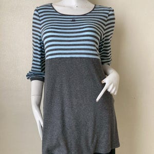 Puede incluir: Una blusa a rayas grises y azules con mangas tres cuartos. La blusa tiene un cuello redondo y una secci&oacute;n inferior gris lisa. Las mangas y la parte superior presentan rayas horizontales en gris y azul claro. La prenda se exhibe en un maniqu&iacute;.