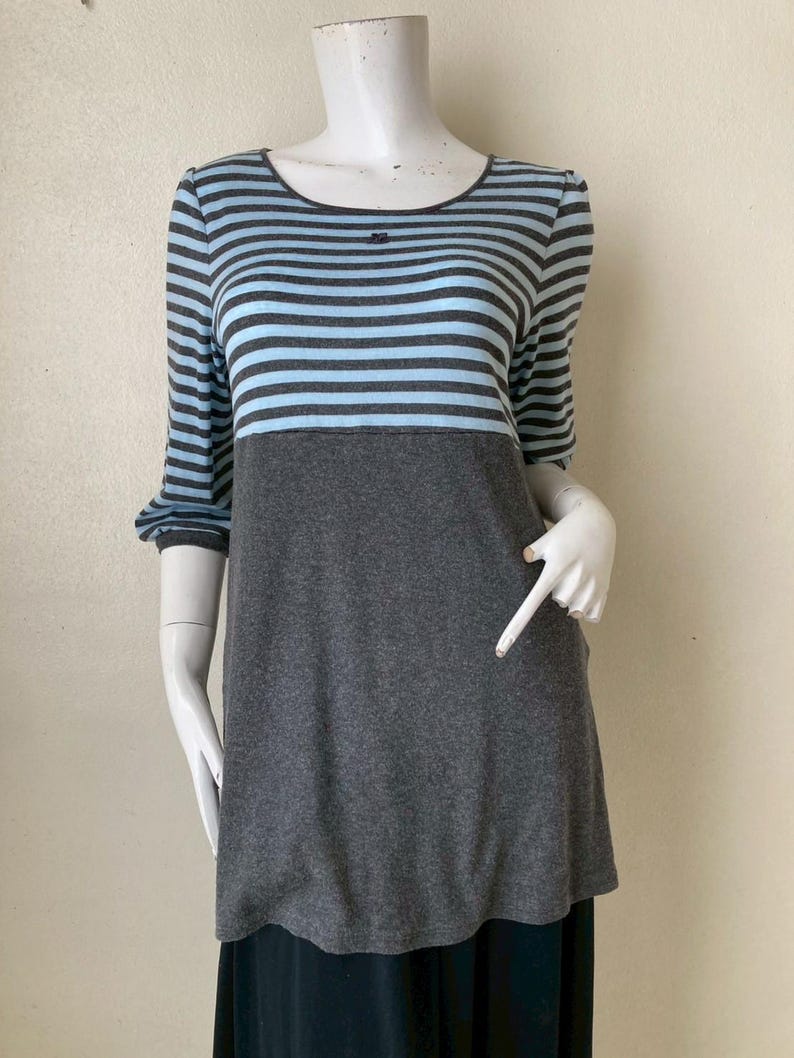 Puede incluir: Una blusa a rayas grises y azules con mangas tres cuartos. La blusa tiene un cuello redondo y una secci&oacute;n inferior gris lisa. Las mangas y la parte superior presentan rayas horizontales en gris y azul claro. La prenda se exhibe en un maniqu&iacute;.