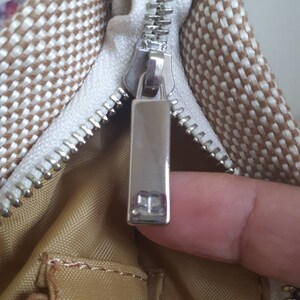 May include: Close-up of a zipper on a brown and white patterned bag. The zipper pull has a silver metal tag with the letter "B" on it.