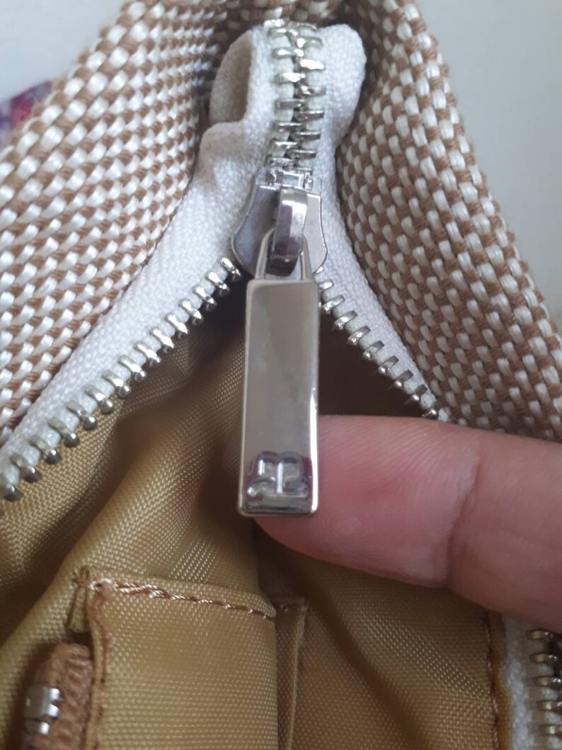 May include: Close-up of a zipper on a brown and white patterned bag. The zipper pull has a silver metal tag with the letter "B" on it.