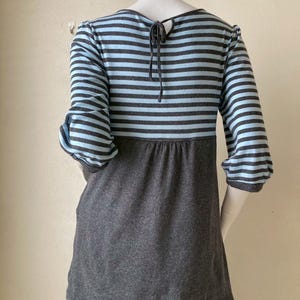 Puede incluir: Una blusa a rayas grises y celeste con mangas tres cuartos. La blusa tiene una secci&oacute;n inferior gris oscuro y un detalle de lazo en la parte posterior del cuello. Las mangas est&aacute;n recogidas en el codo.