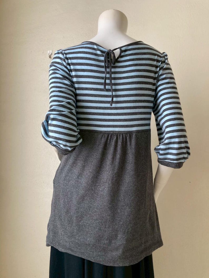 Puede incluir: Una blusa a rayas grises y celeste con mangas tres cuartos. La blusa tiene una secci&oacute;n inferior gris oscuro y un detalle de lazo en la parte posterior del cuello. Las mangas est&aacute;n recogidas en el codo.