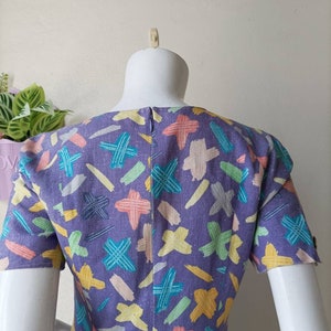 May include: A purple dress with a colorful abstract print of lines and crosses. The dress has short sleeves and a scoop neck.