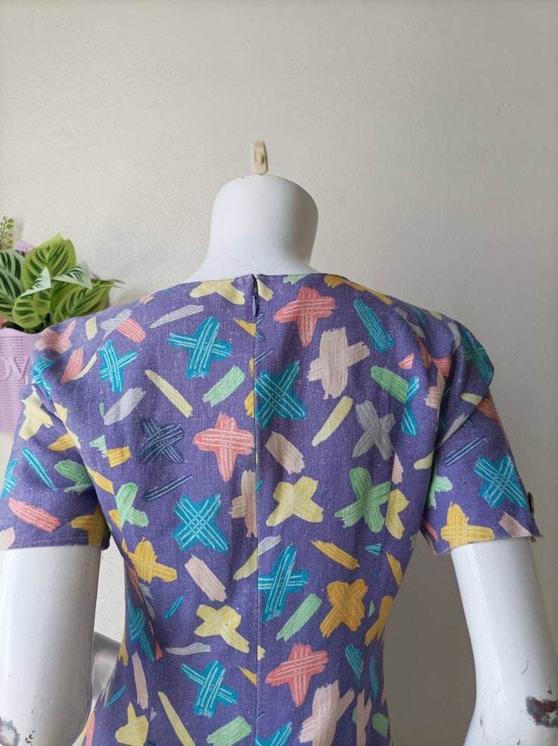May include: A purple dress with a colorful abstract print of lines and crosses. The dress has short sleeves and a scoop neck.